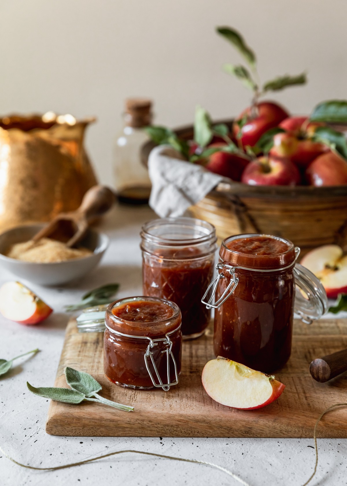 A side image of three jars of sage apple butter on a wood board placed on a grey counter. In the background is a basket of apples, gold teapot, grey bowl of sugar, apple slices, and sage leaves.