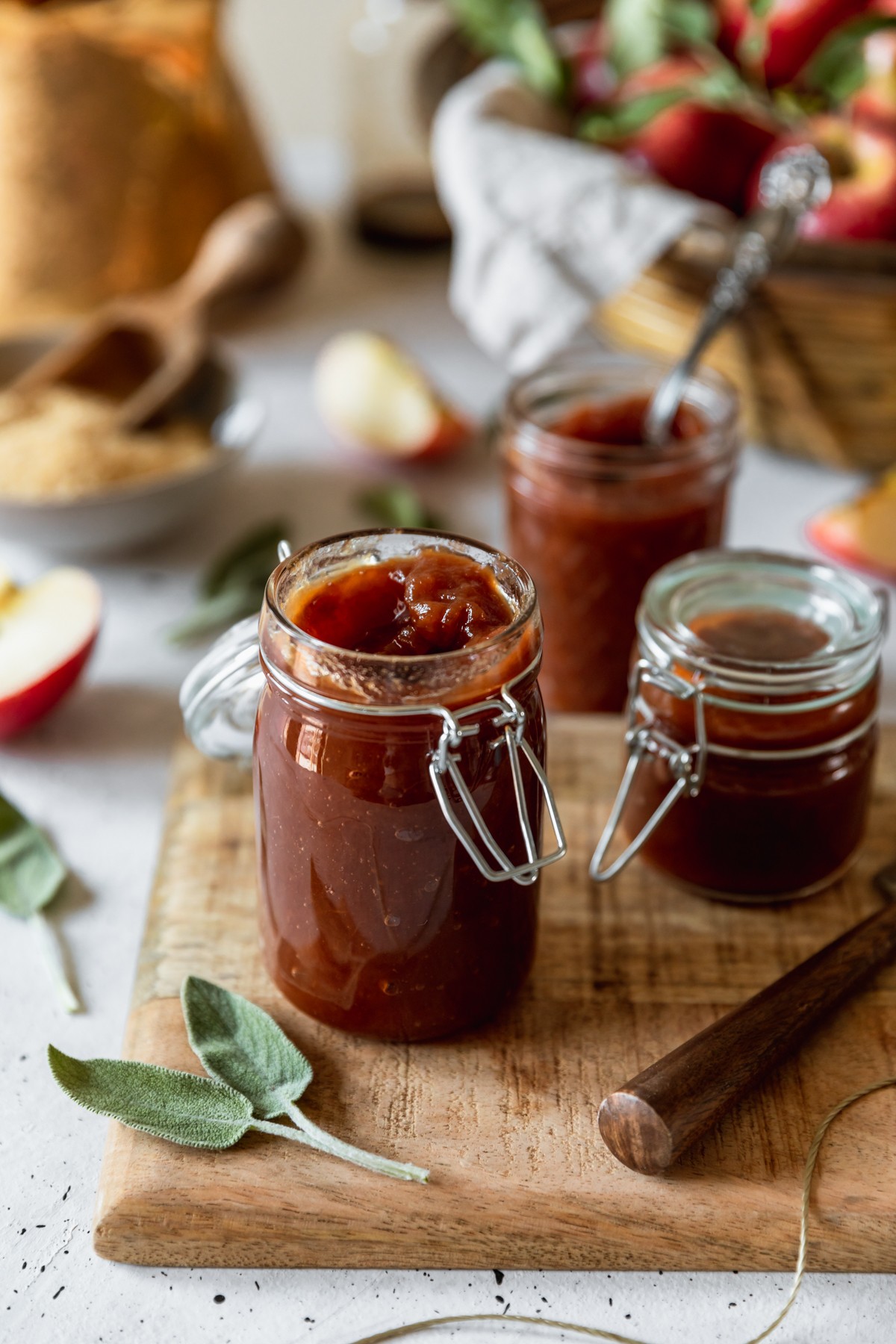 A closeup image of a jar of apple butter on a wood board placed on a white counter. In the background are two more jars of apple butter, a basket of apples, sliced apples, and sage leaves.
