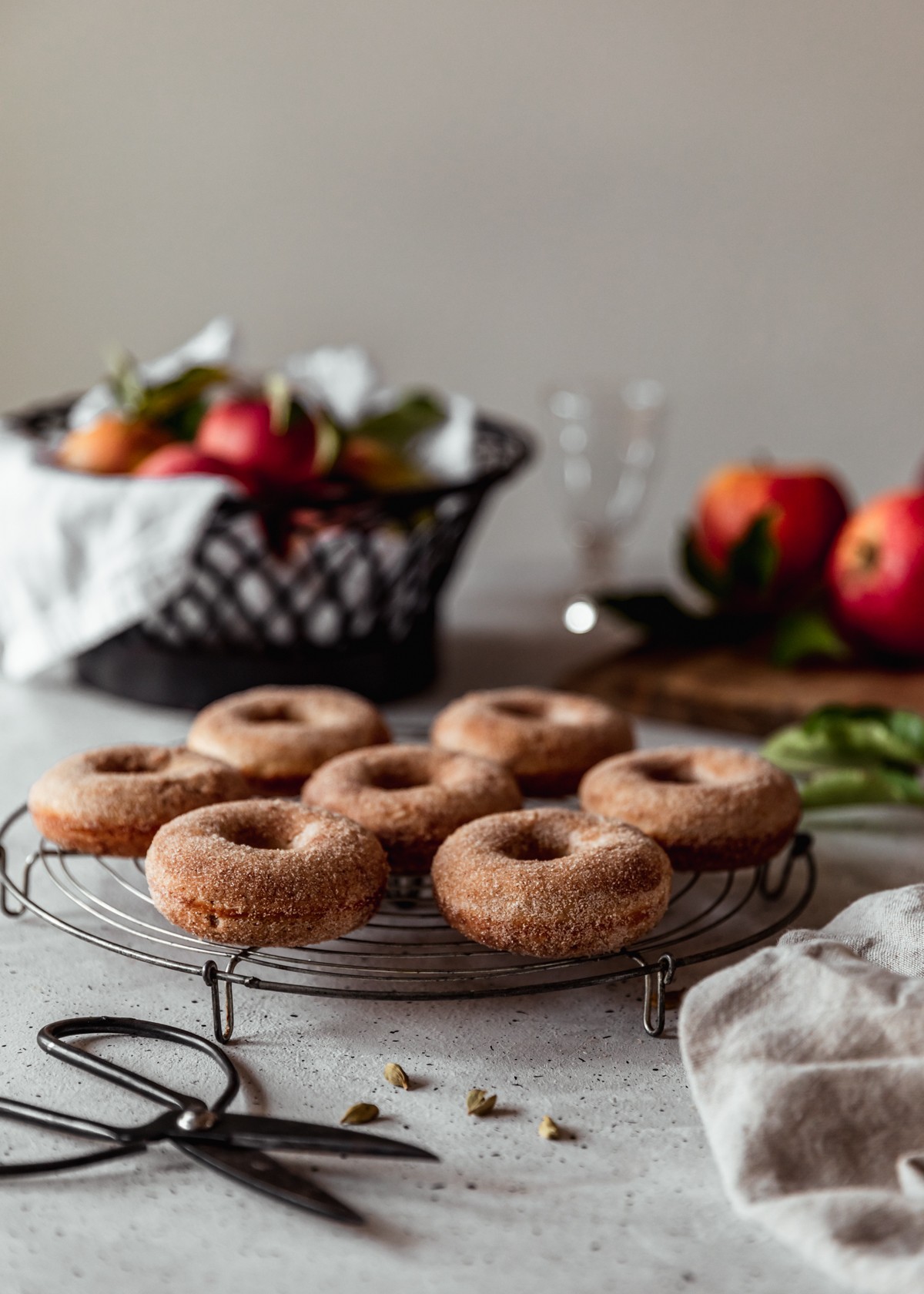 A side image of a circular cooling rack topped with baked apple cider donuts on a white speckled table with a wood tray of apples, black bowl of apples, and beige linen in the background.