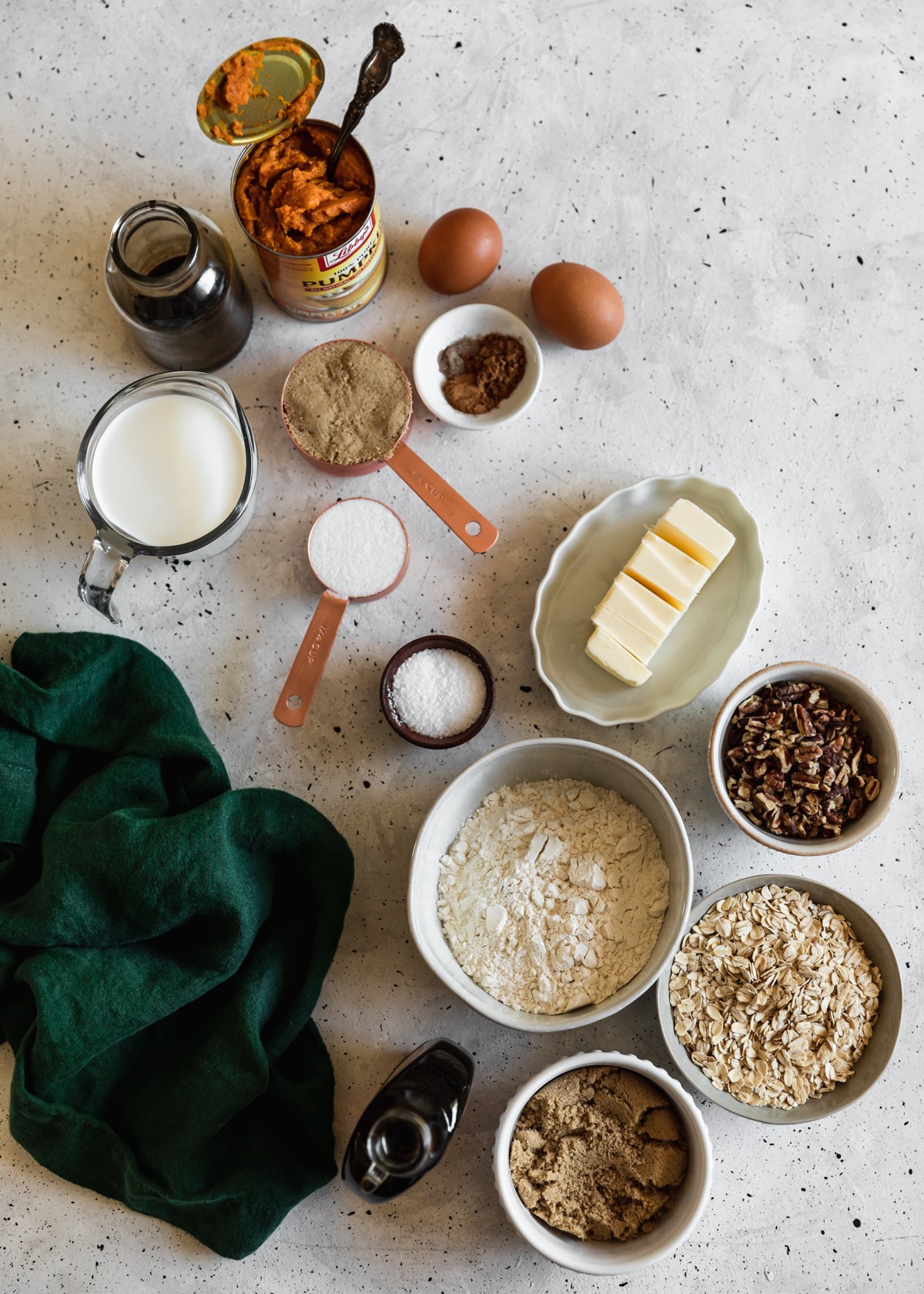 An overhead of various white and grey bowls with baking ingredients like flour, oats, pecans, sugar, and butter on a white speckled counter next to a can of pumpkin and dark green linen.