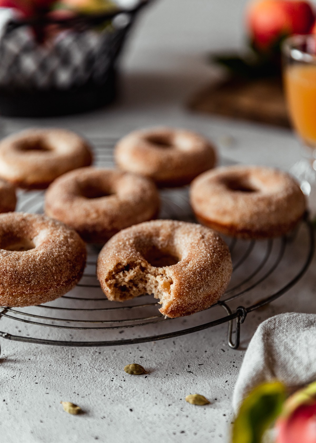 A closeup side image of an apple cider donut with a bite taken out of it on a round cooling rack with more donuts behind it. The cooling rack is on a white speckled table with a black bowl, wood board of apples, and glass of apple cider in the background.
