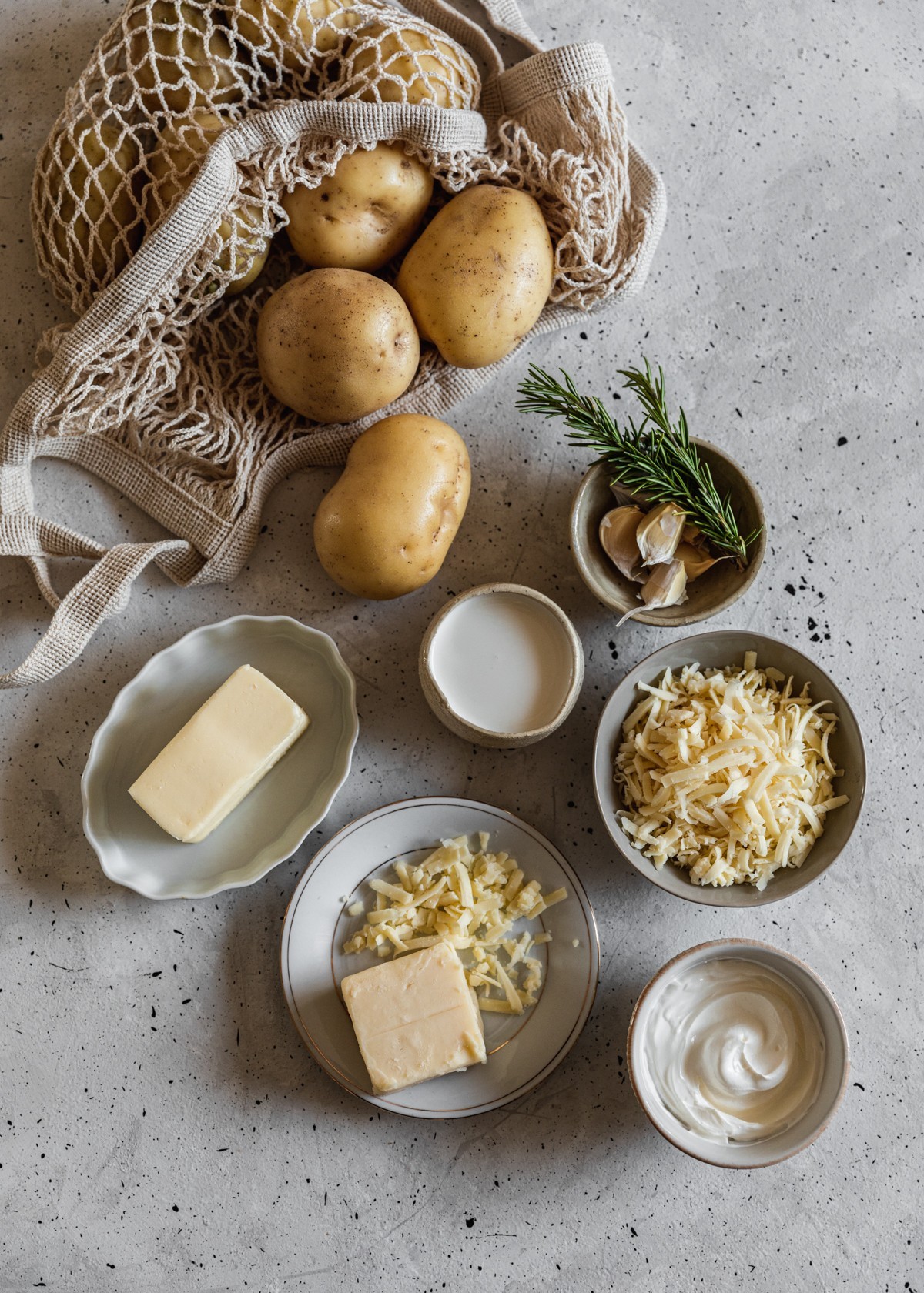 An overhead image of ingredients for cheddar mashed potatoes: a cloth bag of Yukon gold potatoes, a grey bowl of cheese, a white bowl with garlic and rosemary, a white plate with butter, and a white bowl of creme fraiche on a grey table.