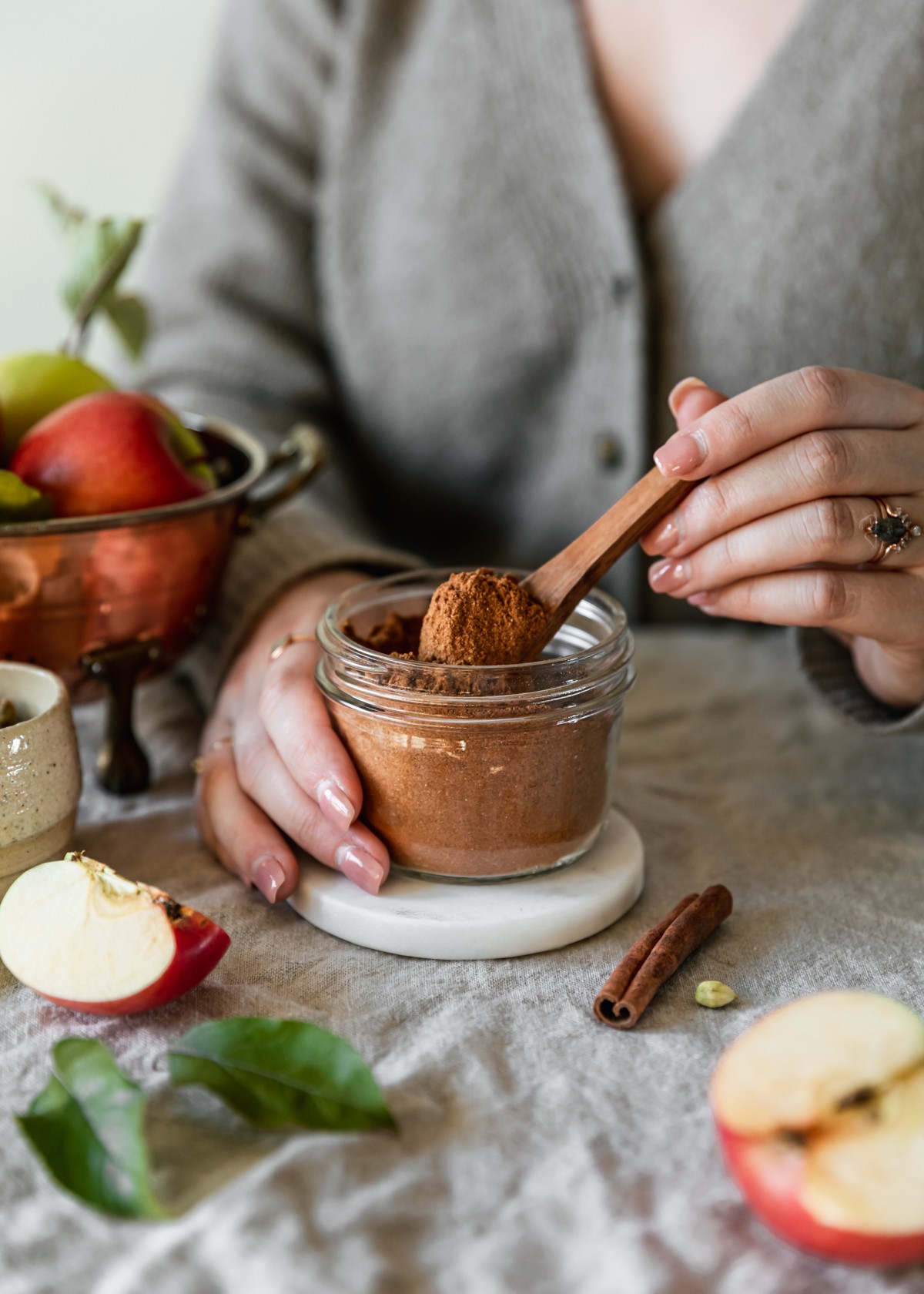 A side image of a woman wearing a beige sweater spooning homemade apple pie spice out of a jar with a wood spoon. The jar is on a beige linen next to a copper colander with apples, sliced apples, and a cinnamon stick.