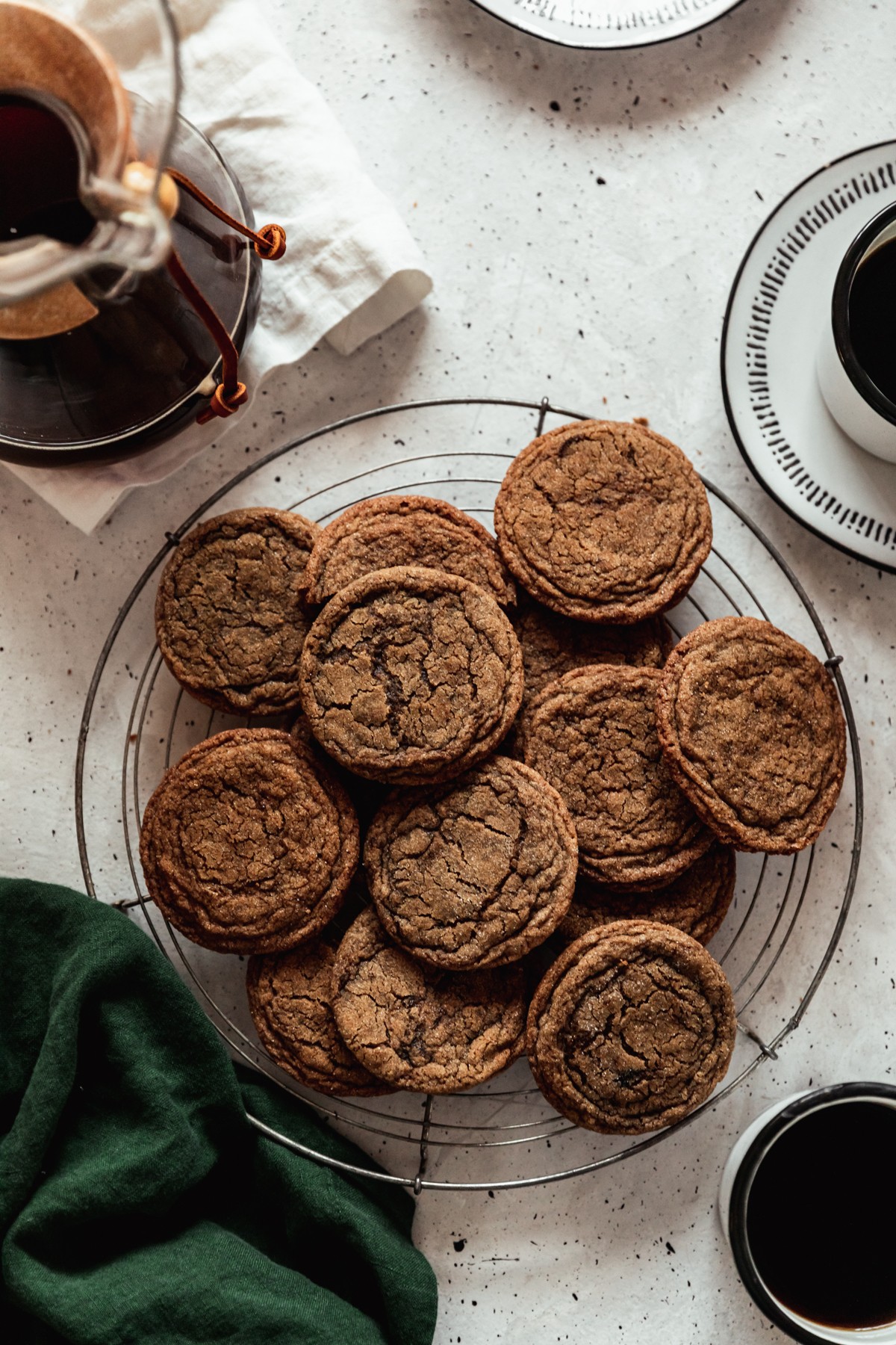 An overhead image of a round cooling rack topped with coffee cookies on a grey speckled table next to a dark green linen, cup of coffee, and Chemex.