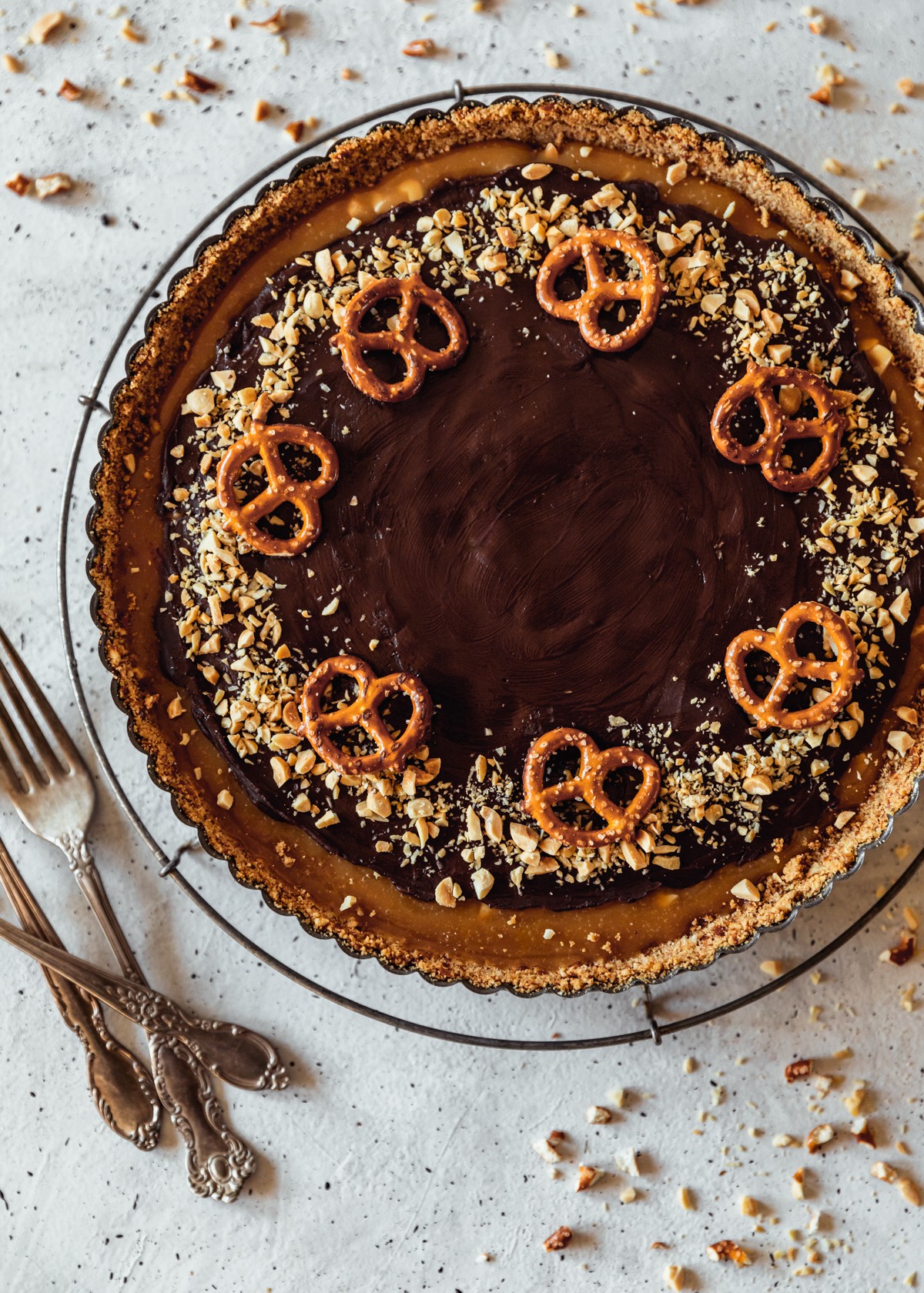 An closeup overhead image of a chocolate caramel tart decorated with chopped peanuts and pretzels. The tart is on a vintage metal cooling rack on a white speckled counter next to vintage forks and chopped peanuts.