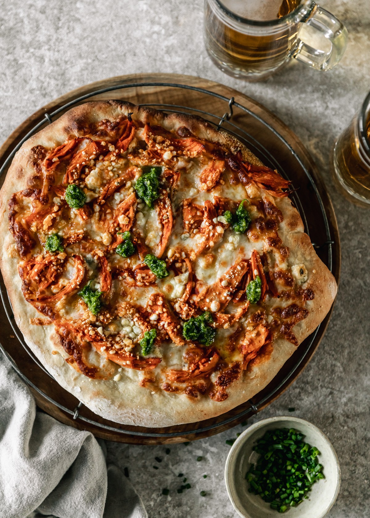 An overhead image of a buffalo chicken pizza with bleu cheese and chive pesto on a vintage cooling rack on a circle wood board placed on a beige table next to a tan linen, white bowl of chives, and cups of beer.