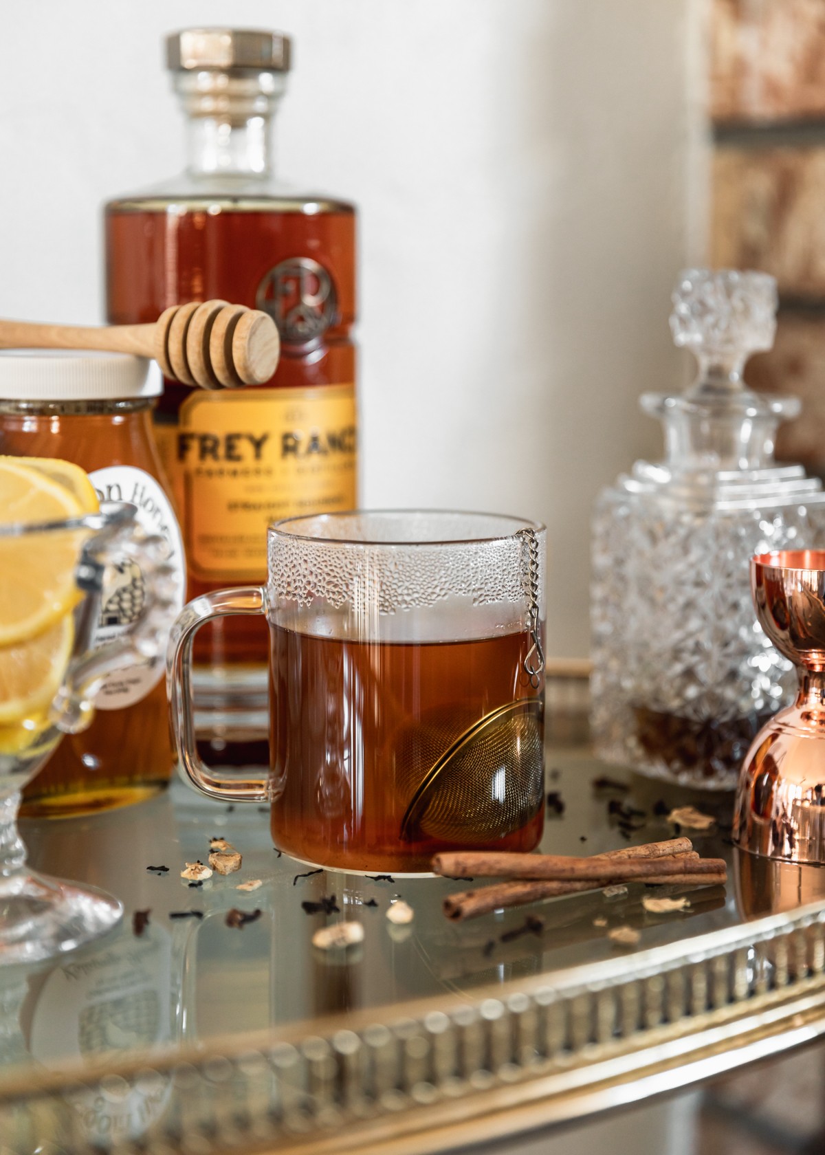 A side image of a cup of black tea in a glass mug on a bar cart next to a bottle of bourbon, jar of honey, white glass of lemon slices, shot, glass, and cinnamon sticks.