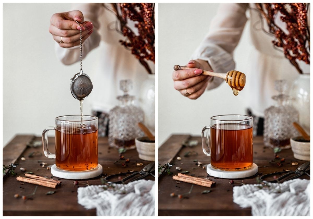 Two side images; on the left, a woman wearing a sheer white shirt is dipping a tea infuser into a glass mug on a wood table next to a white linen, decanter of bourbon, spices, and a white vase of red berry branches. In the right image, the woman is drizzling honey into the mug with a honey stick.