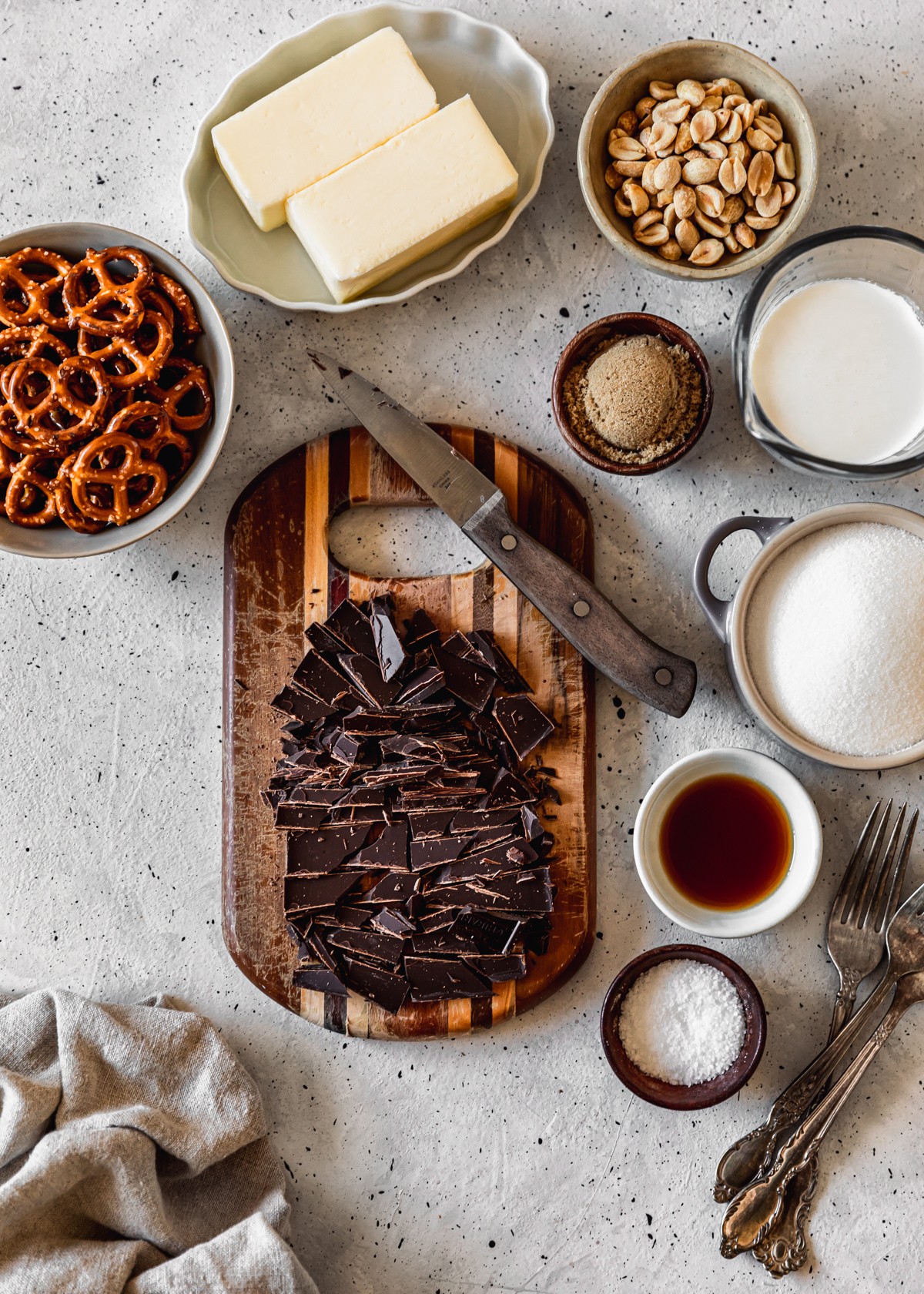 A wood cutting board topped with chopped chocolate on a white and grey speckled counter next to white and cream bowls with sugar, vanilla, salt, butter, pretzels, and peanuts.