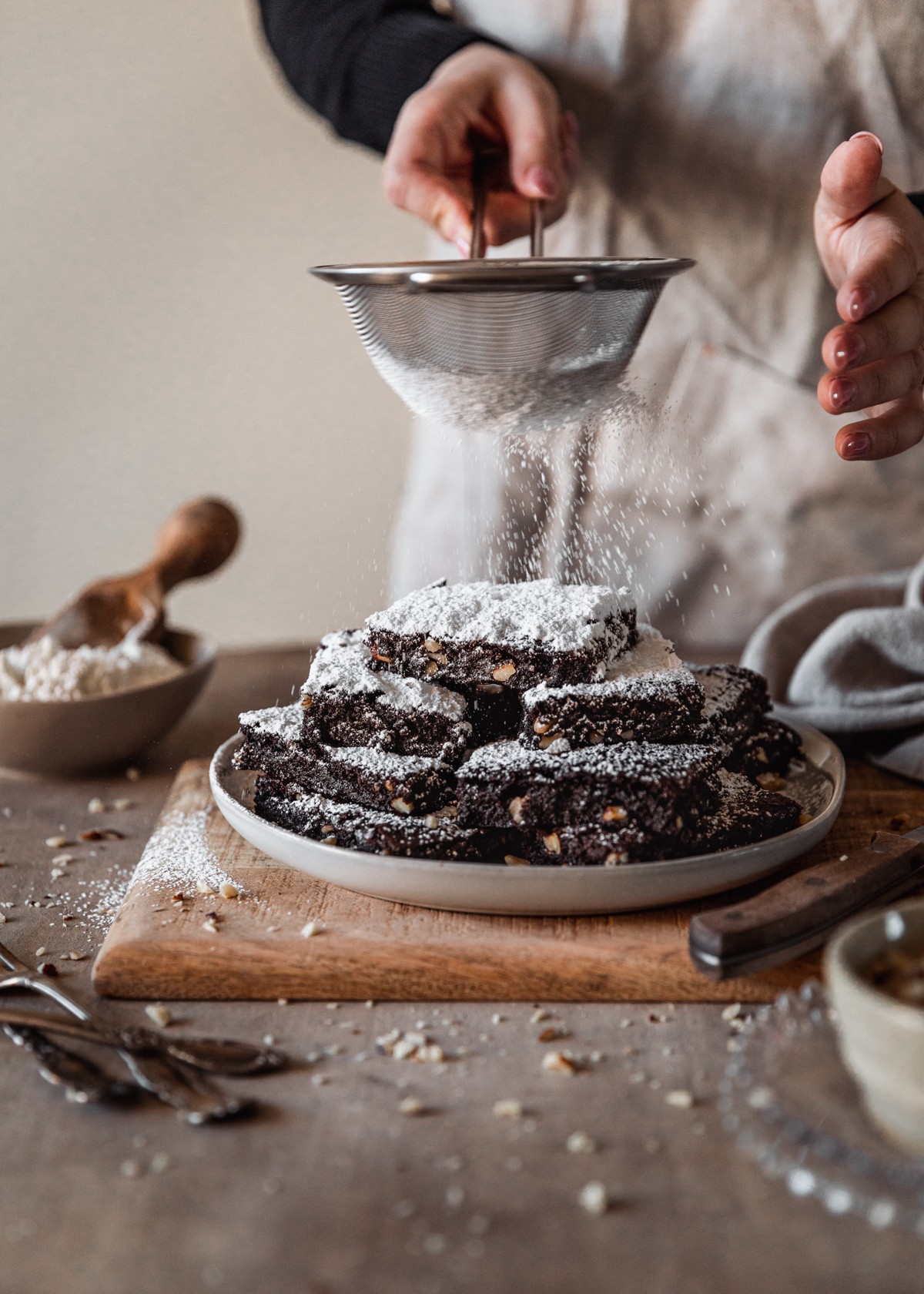A side image of a white plate of 15-minute brownies on a wood board on a brown counter next to a knife, beige linen, and brown bowl of flour. In the background, a woman wearing a beige linen is dusting powdered sugar over the brownies.