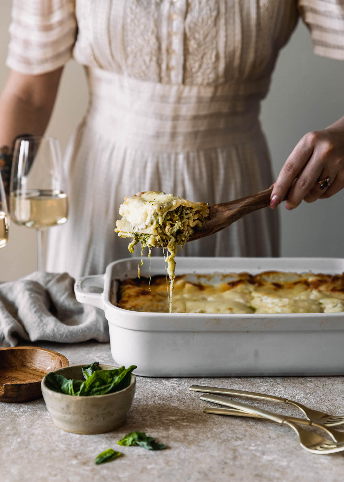 A side image of a woman wearing a white dress spooning white lasagna with pesto and pancetta out of a white casserole dish on a beige table. The lasagna is next to a white bowl of basil, a beige linen, and two glasses of white wine.