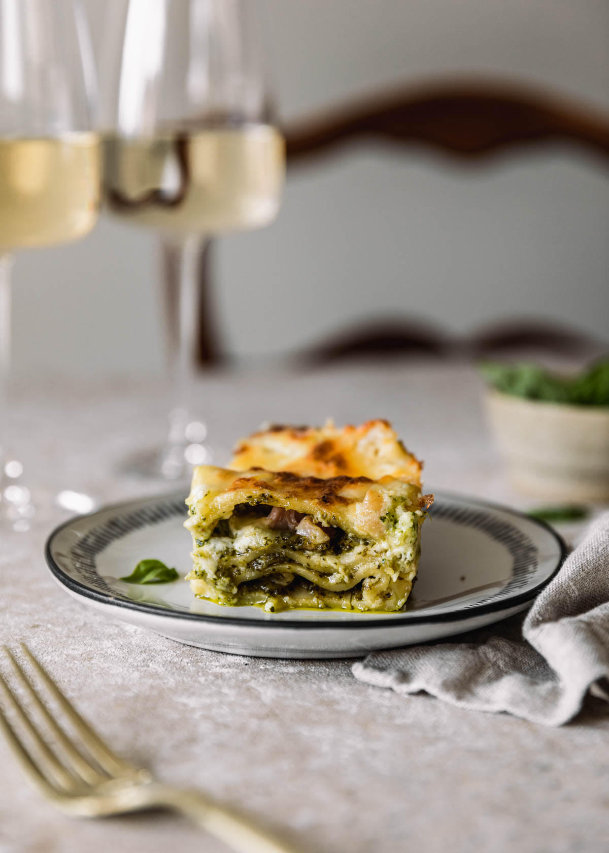 A side image of a white plate with a black rim topped with a slice of pesto white lasagna on a beige table. The plate is next to a beige linen, gold fork, and white bowl of pesto with a wood chair in the background.