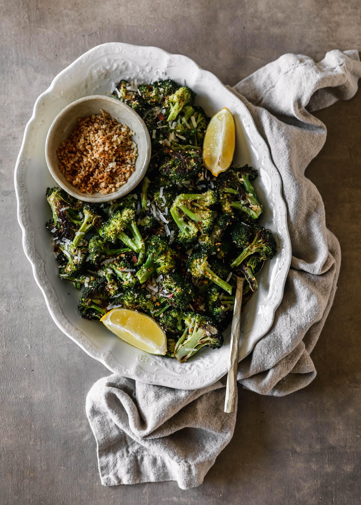 An overhead image of a white dish with charred broccoli on a brown counter next to a beige linen. There is a white bowl of breadcrumbs and lemon wedges on the white dish.