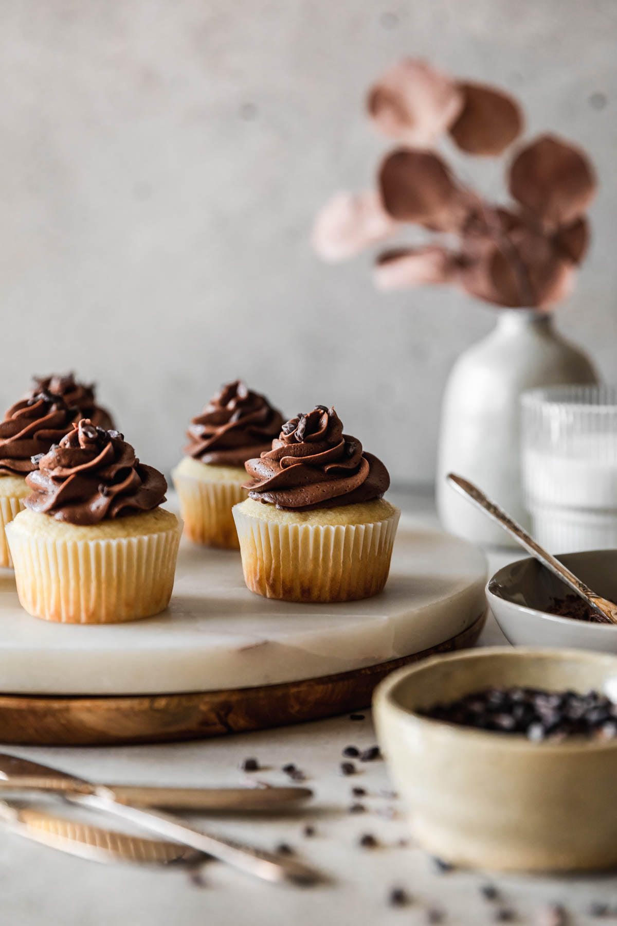 A closeup side image of high altitude yellow cupcakes with chocolate buttercream on a marble and wood cake stand next to a grey vase with blossoms, a glass of milk, a grey bowl of frosting, and a white bowl of cacao nibs with a grey background.