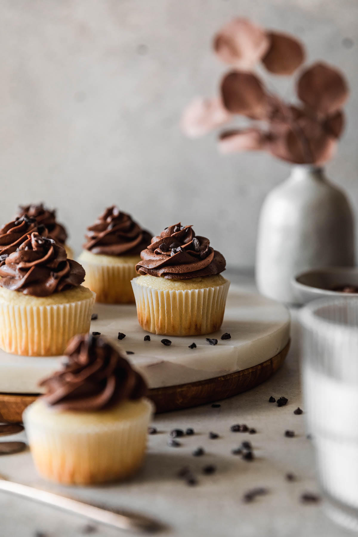 A closeup side image of a marble and wood board topped with high altitude yellow cupcakes with chocolate buttercream on a grey table next to a glass of milk, grey bowl of frosting, and grey vase with dried eucalyptus leaves.