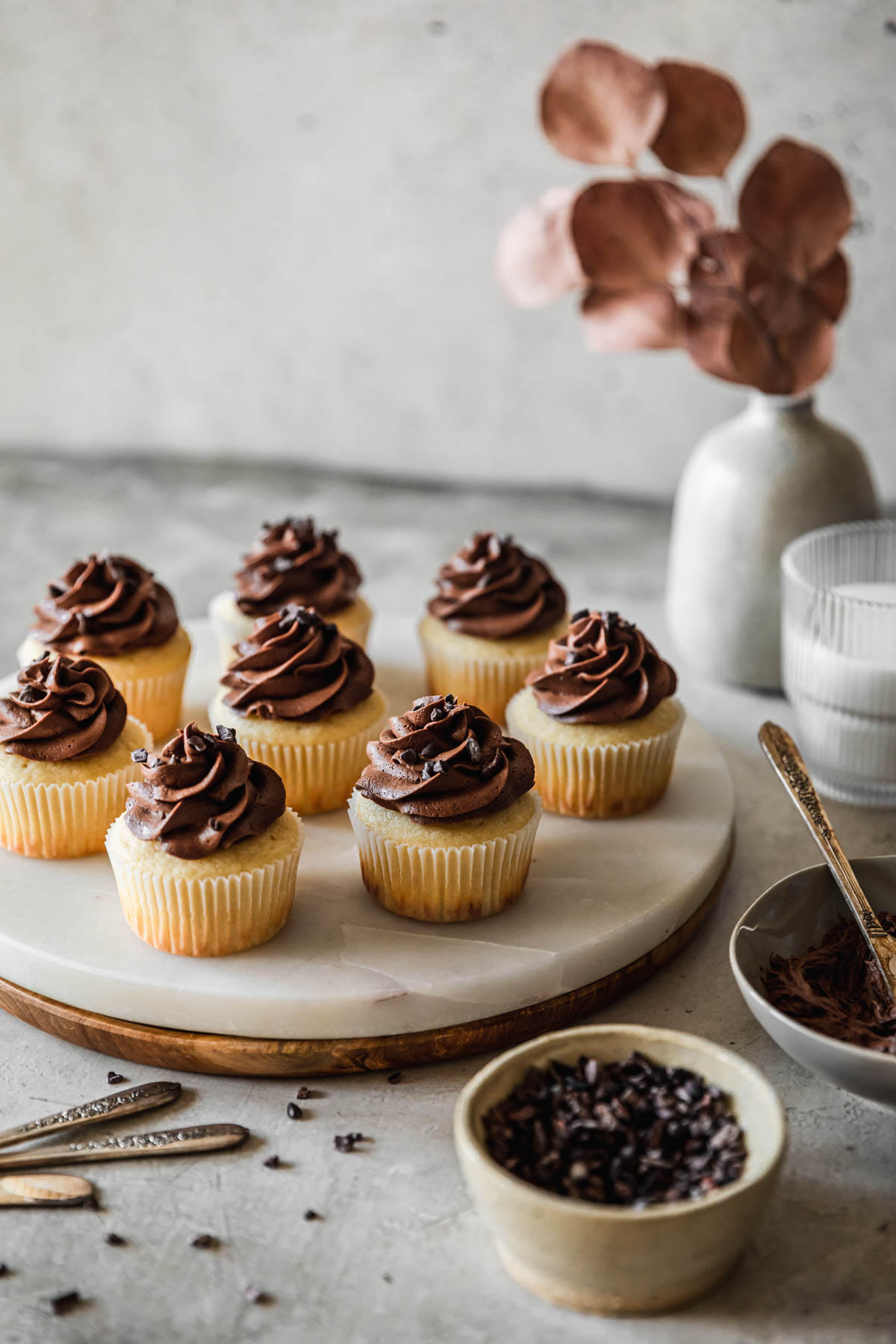A side image of a marble and wood tray of high altitude yellow cupcakes with chocolate buttercream on a grey table next to a grey bowl of frosting, white bowl of cacao nibs, white vase with brown blossoms, and cup of milk.