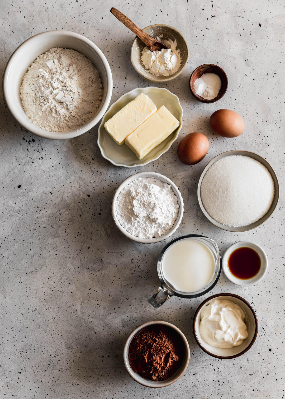 A variety of white and grey bowls filled with baking ingredients like flour, butter, sugar, eggs, cocoa powder, sour cream, and milk on a grey speckled counter.