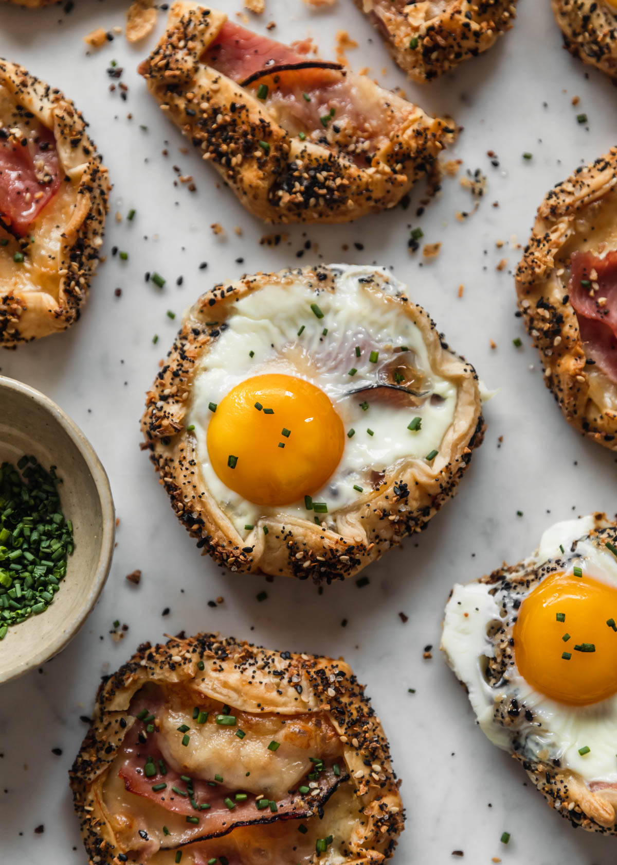 A closeup overhead image of a savory croque madame galette with a runny egg on a white marble counter surrounded by rows of galettes and a white bowl of chives.