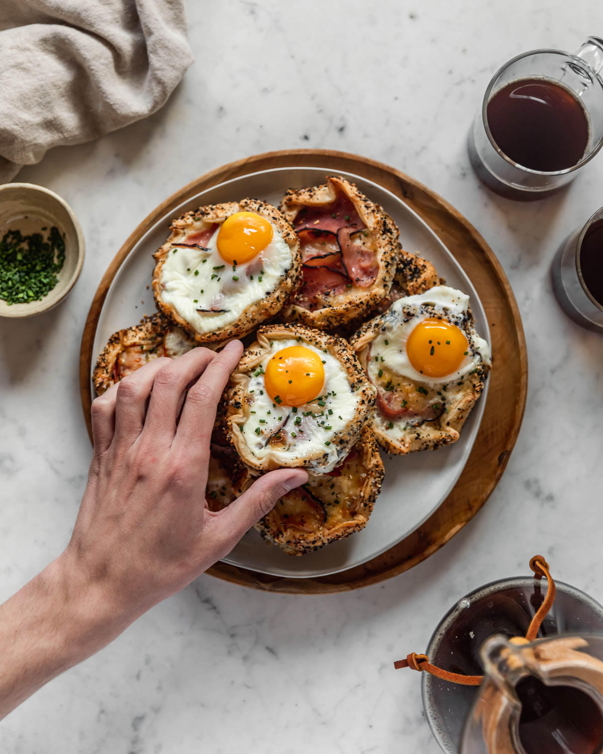 An overhead photo of a man's hand reaching for a croque madame galette on a white plate placed on a wood tray. The tray is next to a Chemex, two cups of coffee, a white bowl of chives, and a tan linen with a marble background.