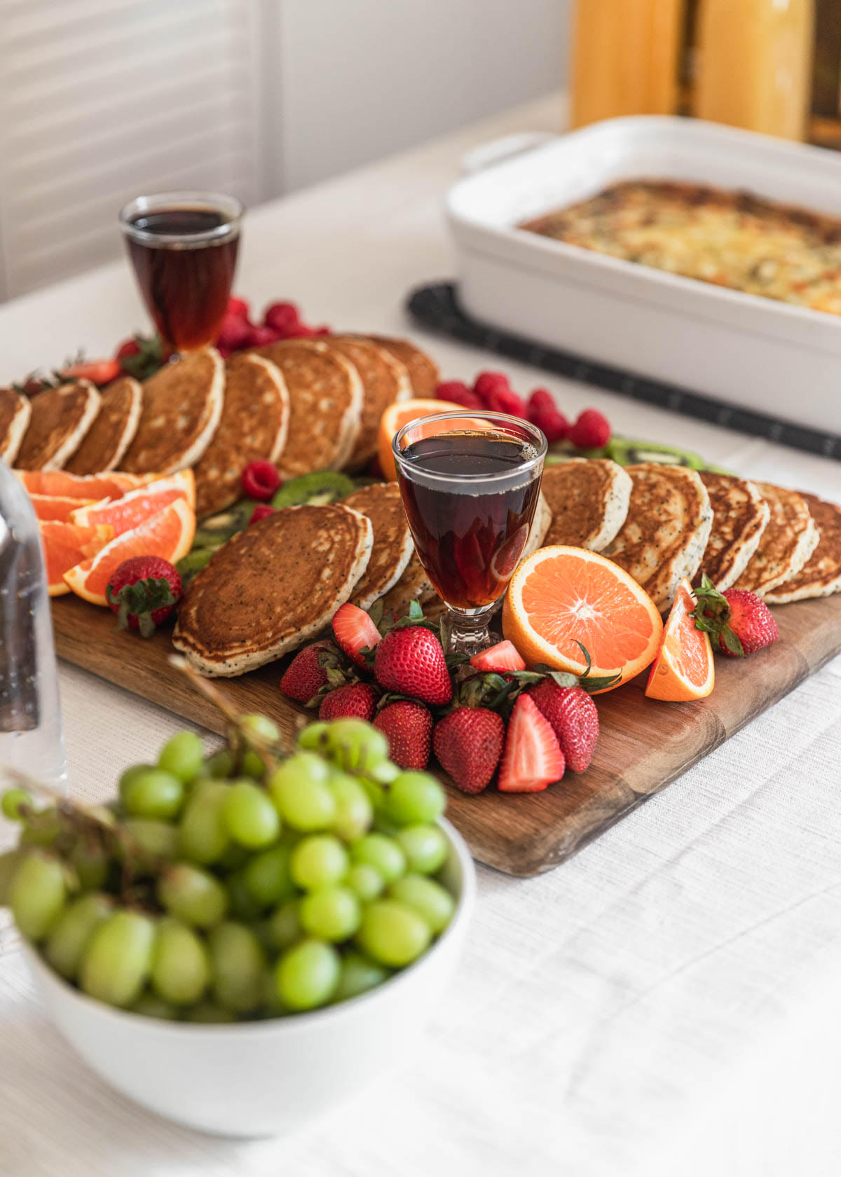 A closeup of a pancake board with fresh fruit and syrup on a white table cloth with breakfast casserole in the background.