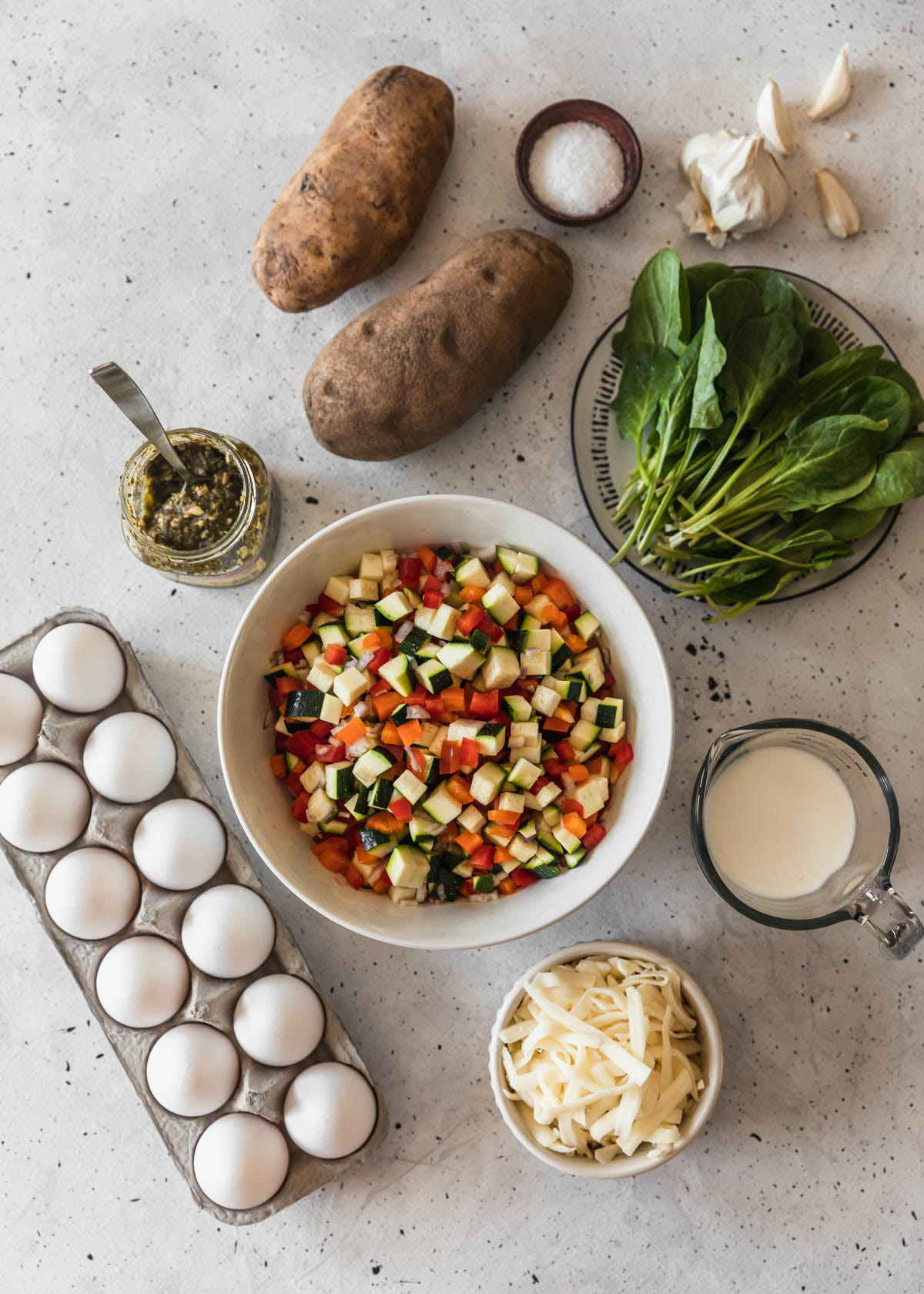 A bowl of chopped veggies on a grey counter next to a white bowl of cheese, eggs, jar of pesto, potatoes, spinach, and a measuring cup of milk.