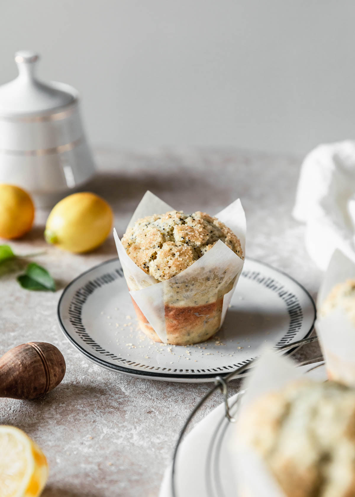 A ricotta lemon poppy seed muffin on a white plate next to a linen and lemons on a tan speckled counter.