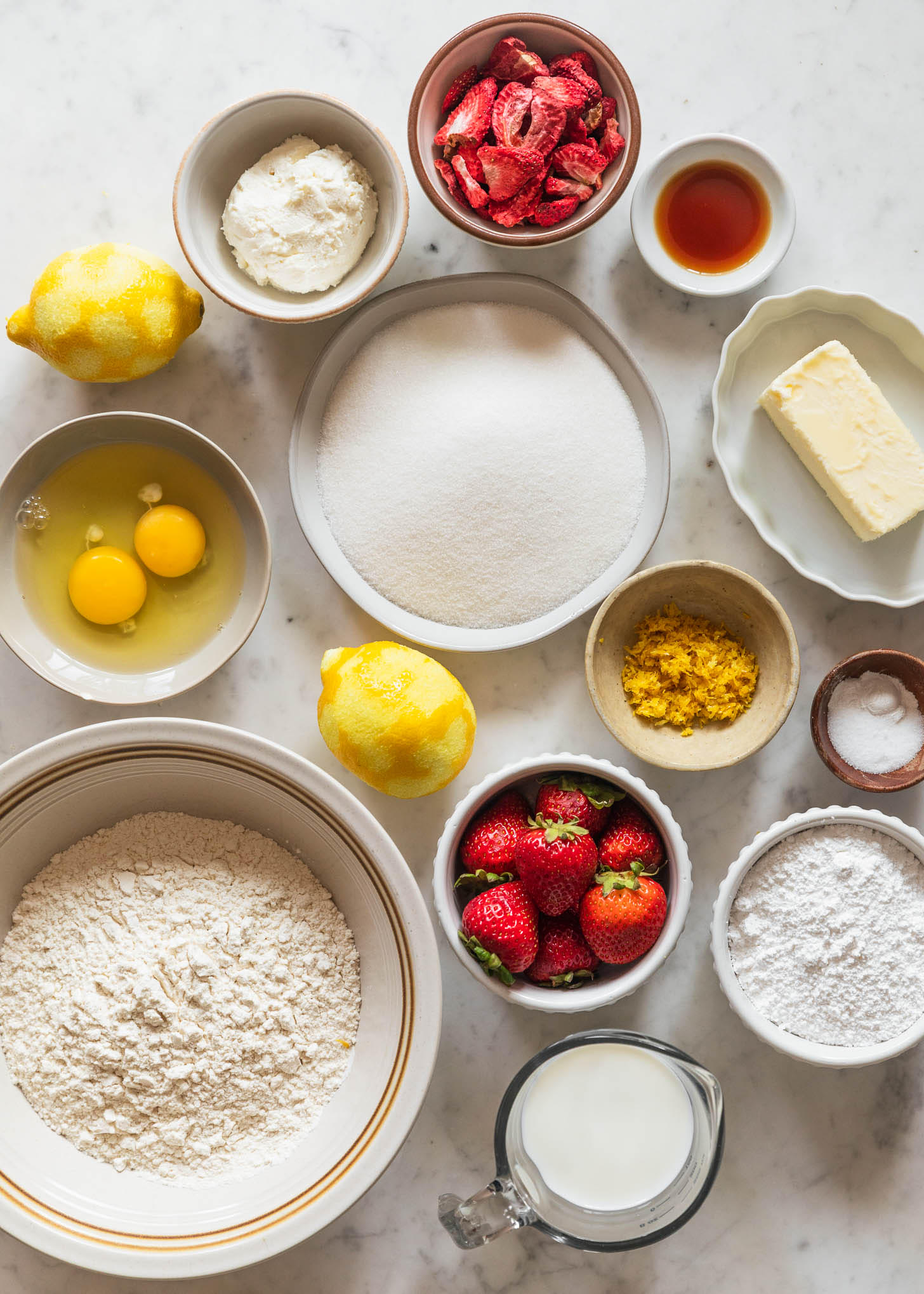 Various white and cream bowls with baking ingredients, strawberries, and lemon zest on a white marble counter.