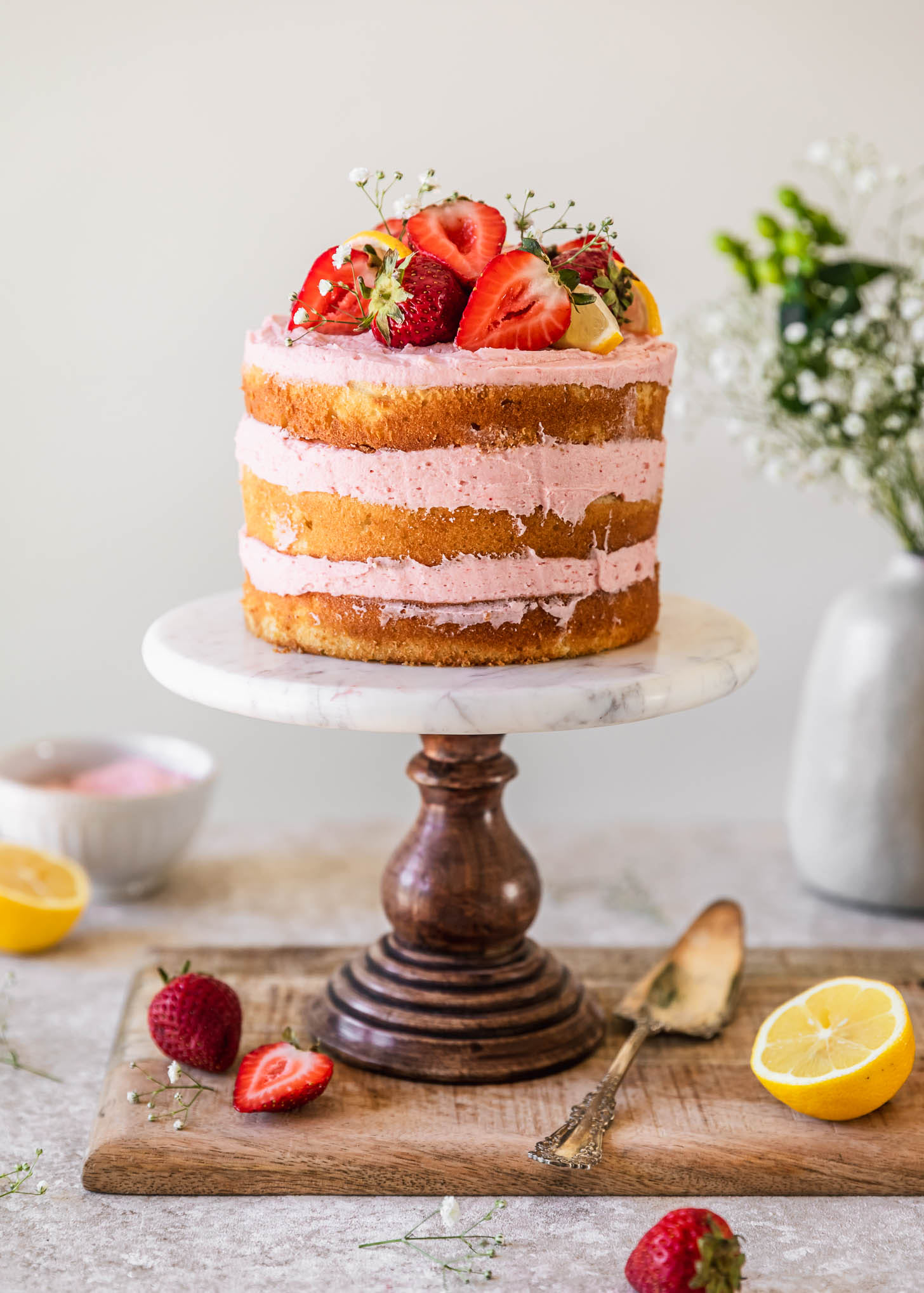 A ricotta strawberry lemon cake on a marble cake stand placed on a wood board next to sliced strawberries, a cake server, and a bouquet of white flowers on a tan counter.